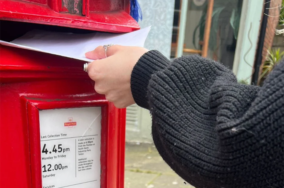 The image shows a person wearing a black sweater posting an envelope into a red Royal Mail postbox. The postbox has a visible collection time notice, indicating the last collection times as 4:45 PM Monday to Friday and 12:00 PM on Saturday. The background includes a blurred view of a building and some plants.
