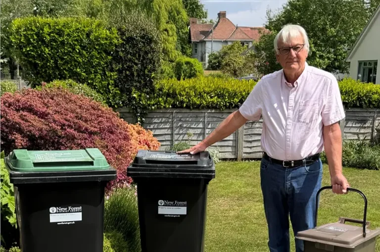 An older man in glasses and a light pink shirt stands in a garden, holding a small brown bin and resting his hand on a black recycling bin—symbolising waste and recycling. Greenery and a house can be seen in the background.
