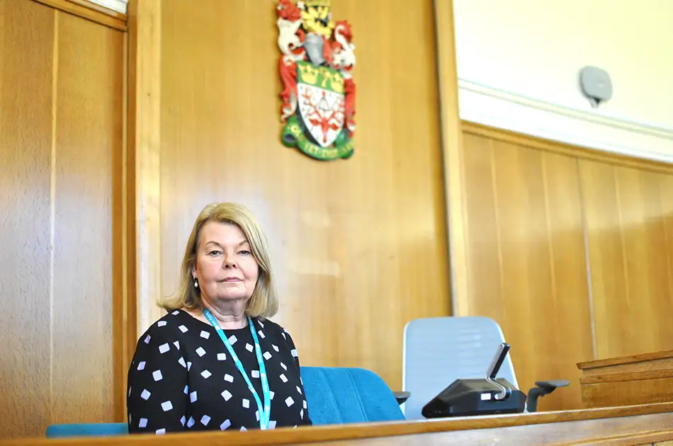 A woman with blonde hair sits in a rural courtroom near a wooden panelled wall, under a colourful coat of arms. She wears a black top with white spots and a teal lanyard, seated next to a microphone on her desk.