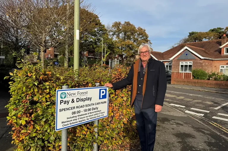 An older man with white hair and glasses stands smiling next to a Pay & Display car park sign on a sunny day, with houses, trees, and a street visible in the background.