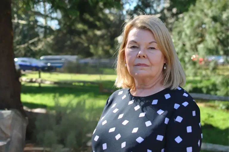 A middle-aged woman with blonde hair, wearing a black top with white geometric shapes, stands outdoors in a sunlit park with green grass and trees in the background.