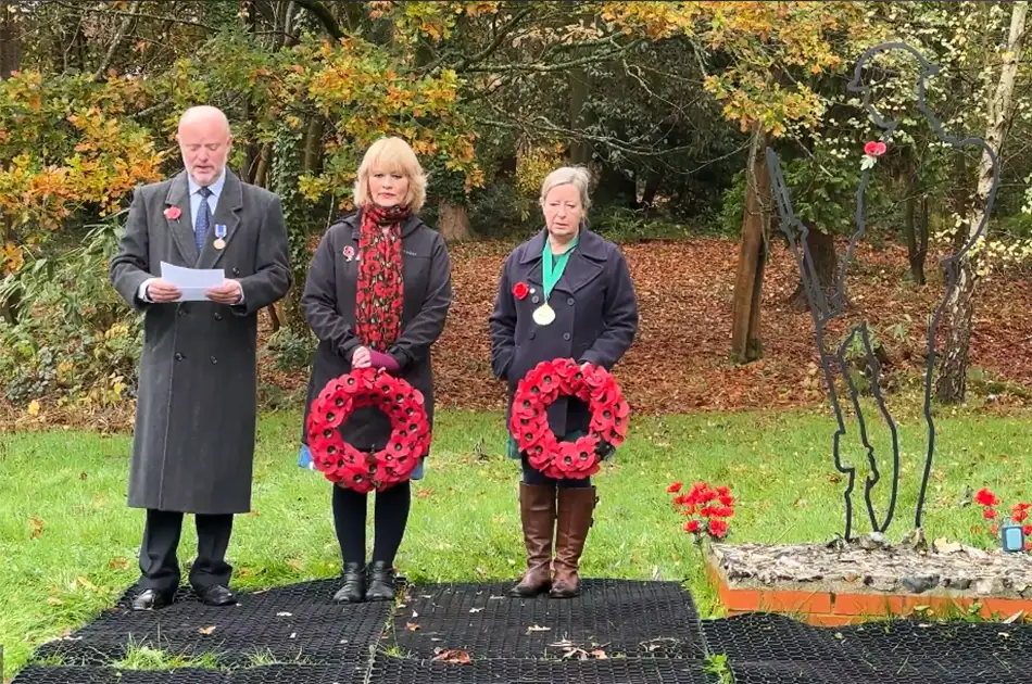 Three people stand by a memorial in a park during autumn, two holding poppy wreaths. A metal soldier silhouette and a flower arrangement are in the foreground; trees with autumn leaves are in the background.
