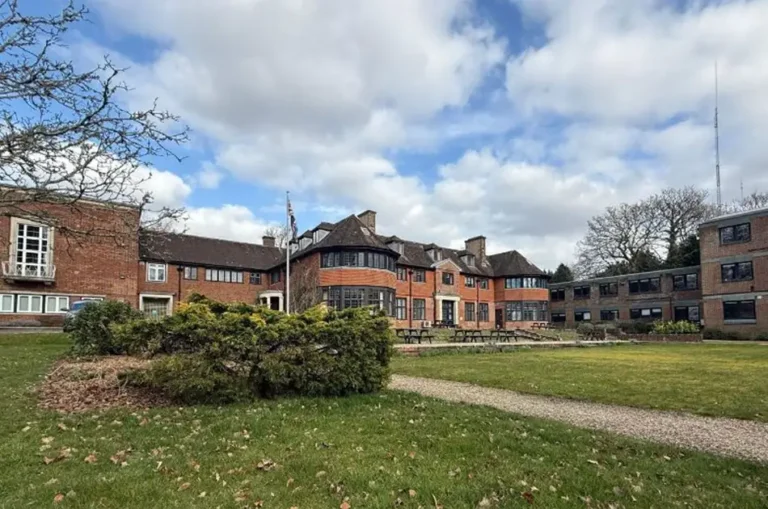 A large brick building with multiple wings and chimneys, possibly linked to local government reorganisation, is set behind a green lawn and hedges under a partly cloudy sky. Outdoor tables and chairs are arranged near the building.