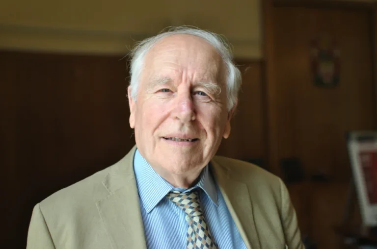 An elderly man with white hair wearing a beige suit jacket, blue striped shirt, and patterned tie, smiles softly whilst standing indoors in a warmly lit room with wooden walls—reflecting the thoughtful spirit of New Forest residents during the Local Plan consultation.