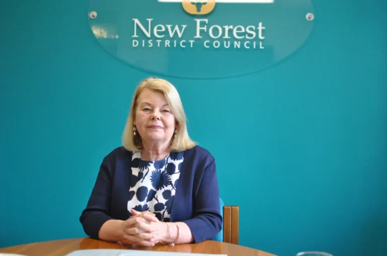 A woman with blonde hair sits at a table with her hands clasped, in front of a teal wall with a sign that reads New Forest District Council. She is wearing a patterned top and a dark blue jacket.