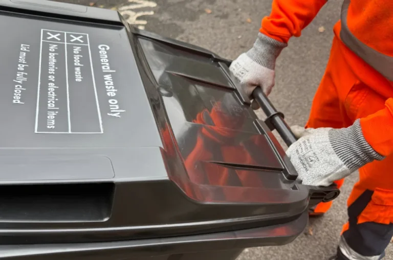 A person wearing orange work clothes and grey gloves is wheeling a black general waste bin with a closed lid along a street during rubbish collections. The bin label lists prohibited items.