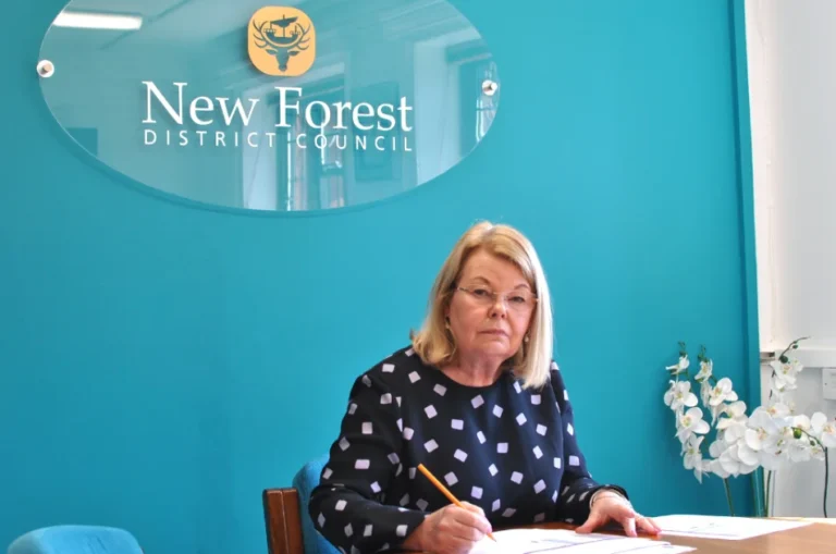 A woman sits at a desk, writing with a pencil, in front of a turquoise wall with an update from the New Forest District Council. White flowers are on the desk beside her.