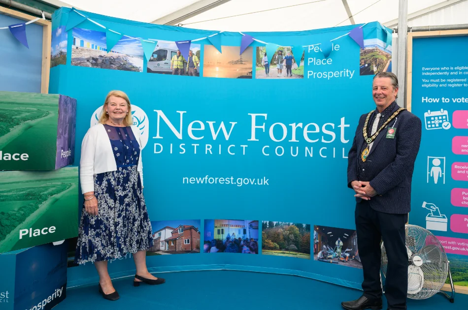 Two officials stand smiling in front of a blue New Forest District Council display, featuring photos of local scenes, large labelled blocks, and a fan on the floor—perhaps highlighting recent local government changes in the New Forest showcased at the Hampshire County Show.