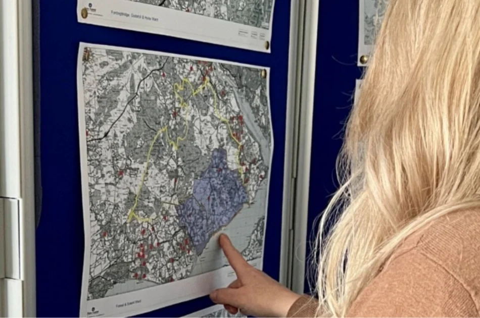 A person with long blonde hair points at a black-and-white map with coloured markings, pinned to a blue board as part of a Local Plan review. Other maps are also visible on the board.