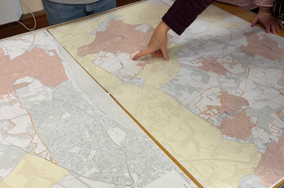 Two people stand over large, detailed maps spread on a table. One person points to a specific area on the map, which shows regions shaded in different colours with outlined boundaries—suggesting the council is considering a new direction for the Local Plan.